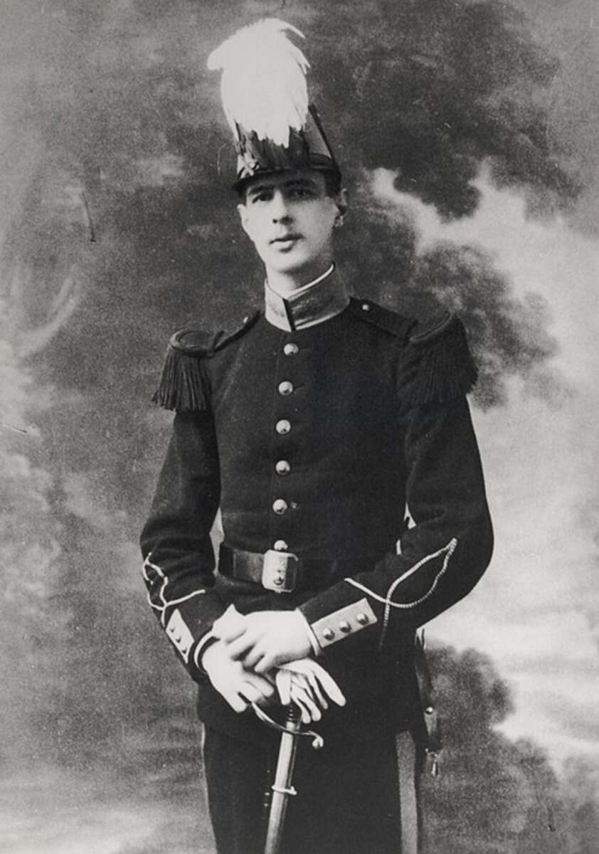 A young man in a formal military uniform and a hat with a feather stands holding a sword, posing for a vintage black-and-white portrait against a painted backdrop.