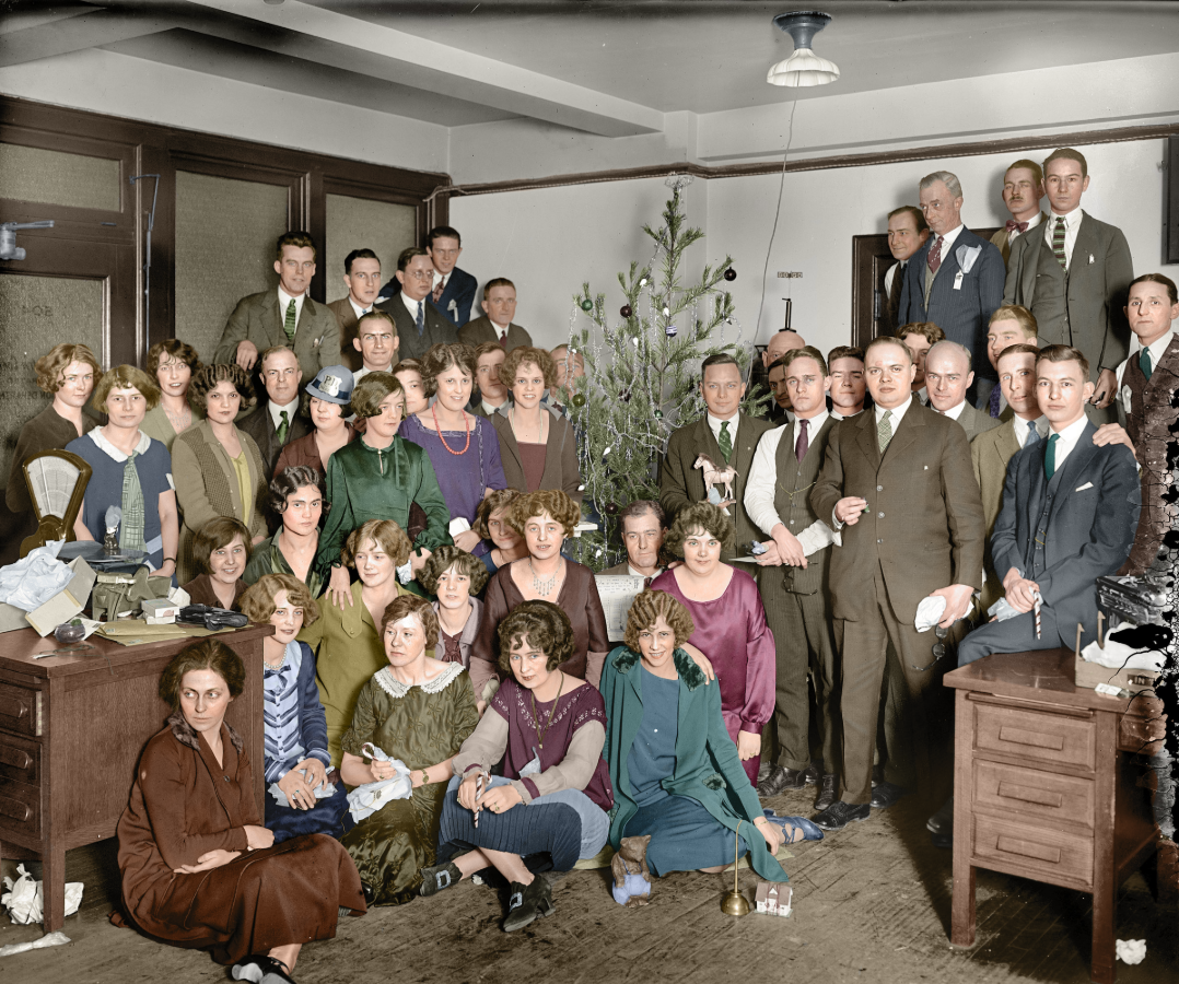 A large group of men and women in vintage clothing gather around a small decorated Christmas tree in an office, posing for a group photo. Some people are seated while others stand around desks.