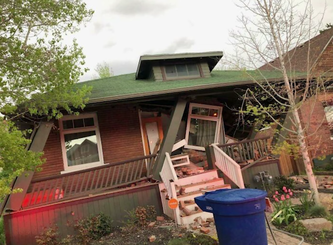 A brick house with a green roof is heavily damaged and leaning to one side. The front porch and steps are collapsed, debris is scattered, and a blue recycle bin sits at the curb. Trees and a flower bed are visible nearby.