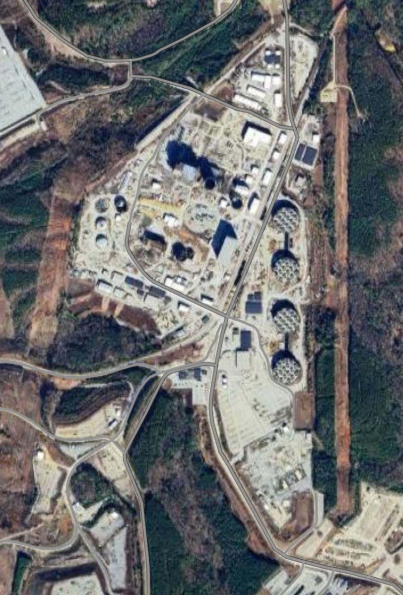 Aerial view of an industrial facility surrounded by trees, featuring several large circular and rectangular structures, roads, and buildings spread across the complex.