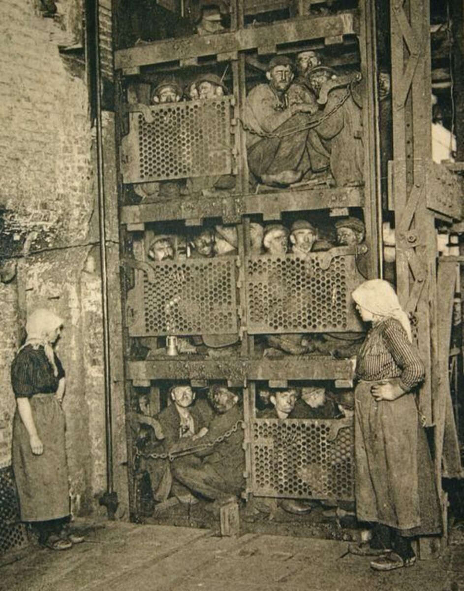 Black-and-white photo of miners packed tightly into multi-level industrial elevator cages, with two women standing outside, likely supervisors or workers, at a mining site. The miners appear tired and crowded.