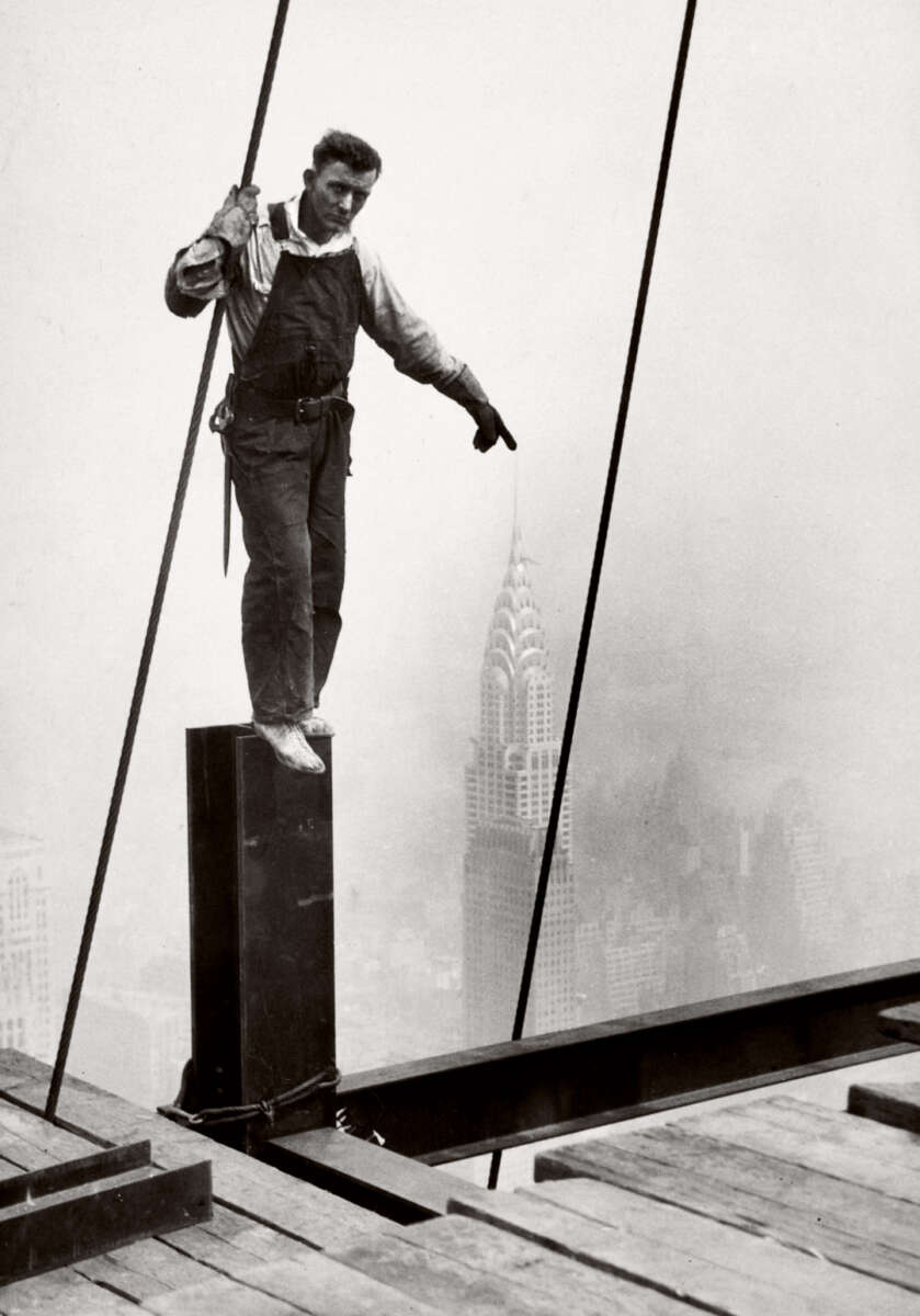 A construction worker balances on a steel beam high above New York City, with the Chrysler Building visible in the foggy background. He is wearing overalls and holding onto a cable for support.