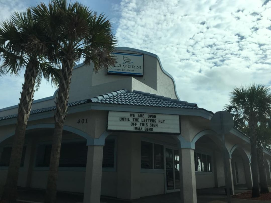 A white and blue restaurant with palm trees out front. The sign reads, "WE ARE OPEN UNTIL THE LETTERS FLY OFF THIS SIGN IRMA GERD," referencing Hurricane Irma. The sky is partly cloudy.