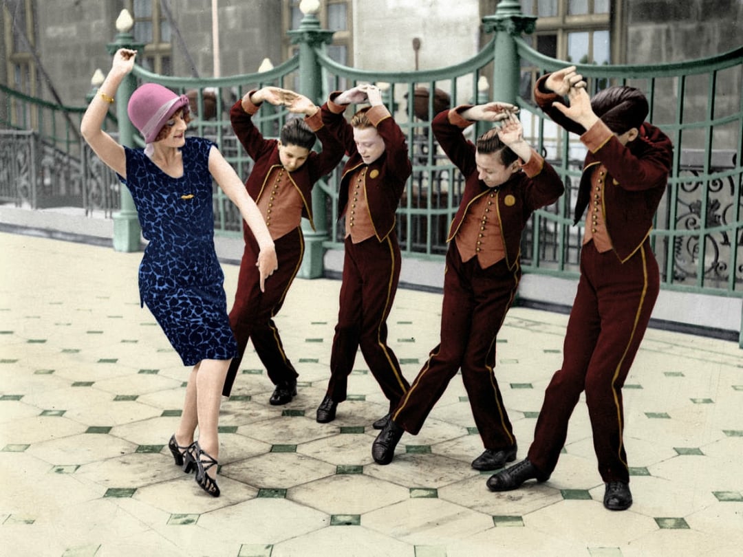 A woman in a blue dress and pink hat dances playfully, while four boys in matching brown uniforms pose behind her with arms raised, mimicking her movements on a tiled outdoor patio.