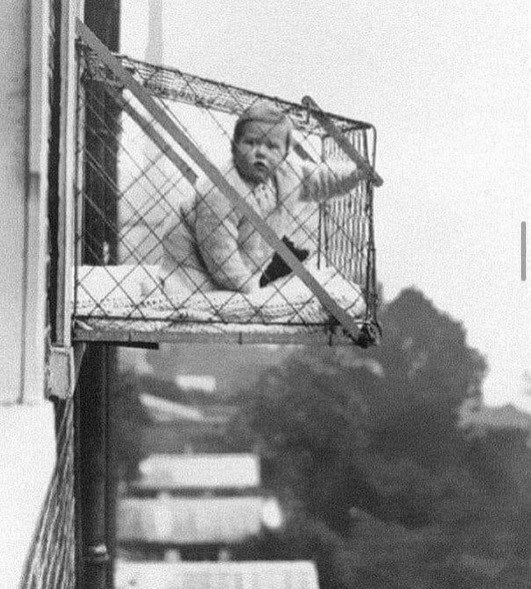 A baby sits in a wire mesh cage attached to the outside of an apartment window, high above the ground. The cage extends outward, and the baby looks toward the camera. Trees and rooftops are visible below.