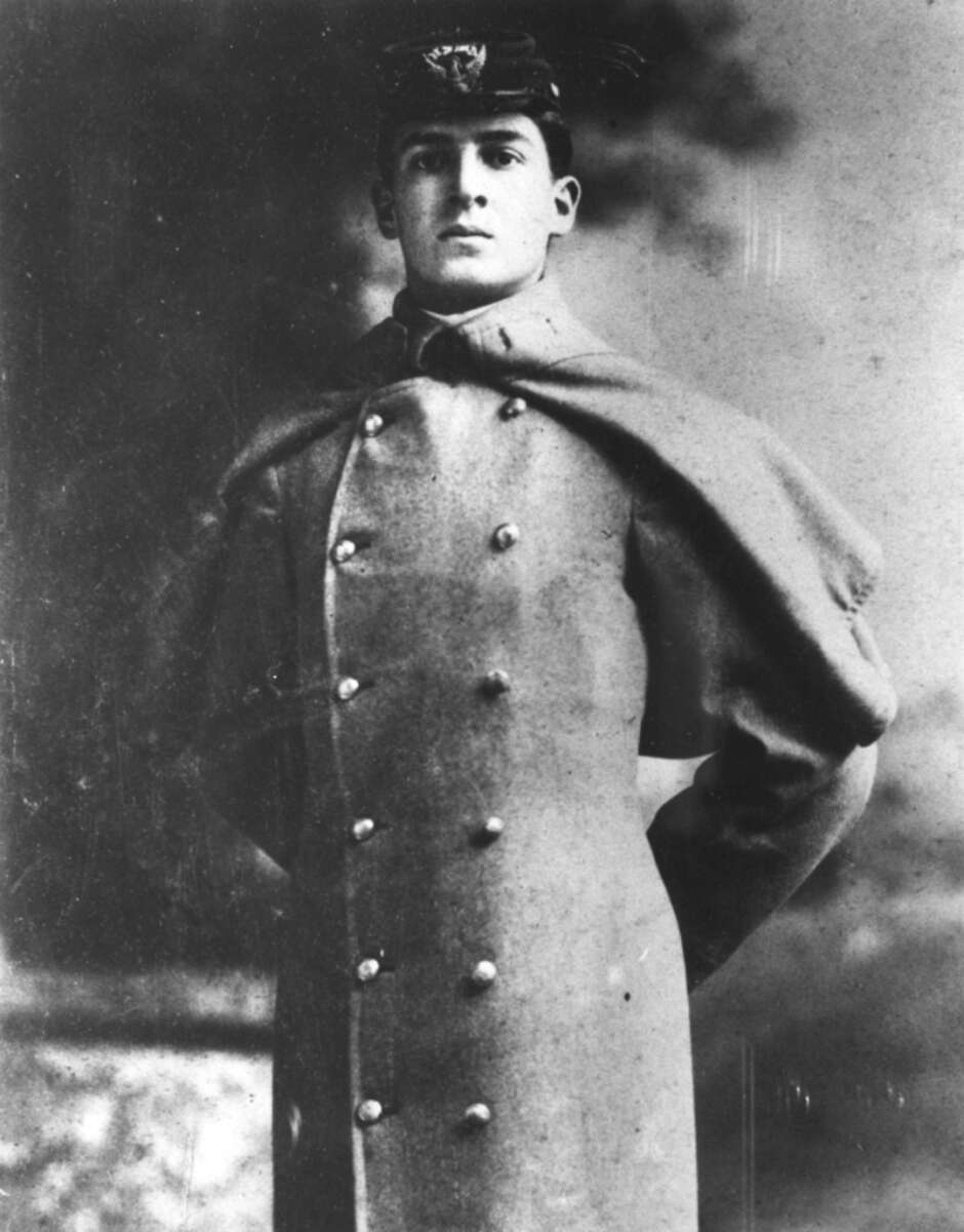 A young man in a military-style coat and cap stands with his hands behind his back, looking confidently at the camera. The black-and-white photo has a formal and historic appearance.