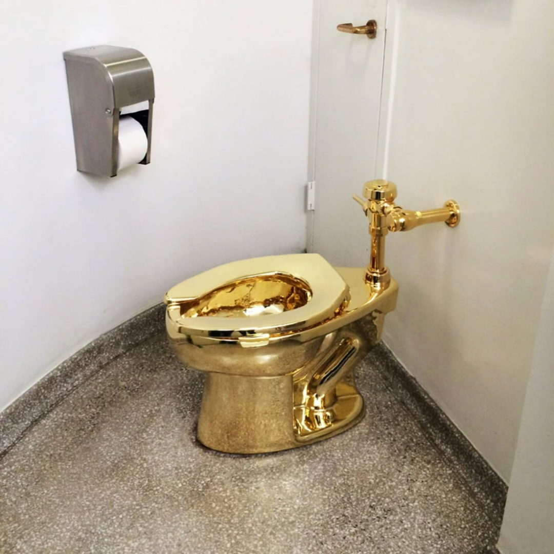 A shiny gold toilet is installed in the corner of a bathroom with white walls, a metal toilet paper dispenser, and speckled gray flooring.