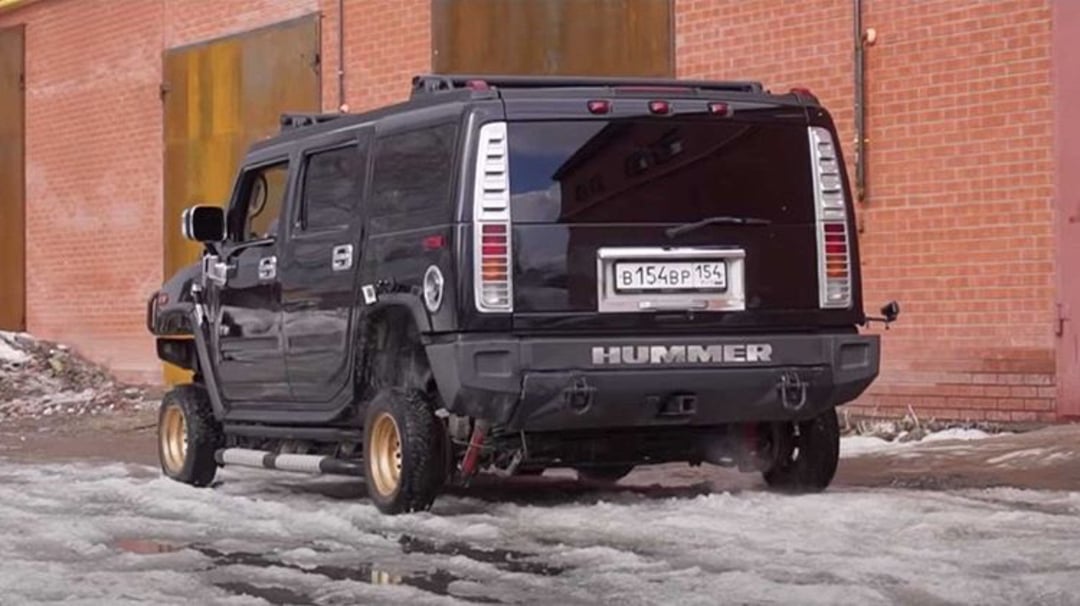 A black Hummer SUV with unusually small, thin wheels is parked on a snowy surface near a red brick building. The vehicle’s large frame contrasts with its tiny wheels.