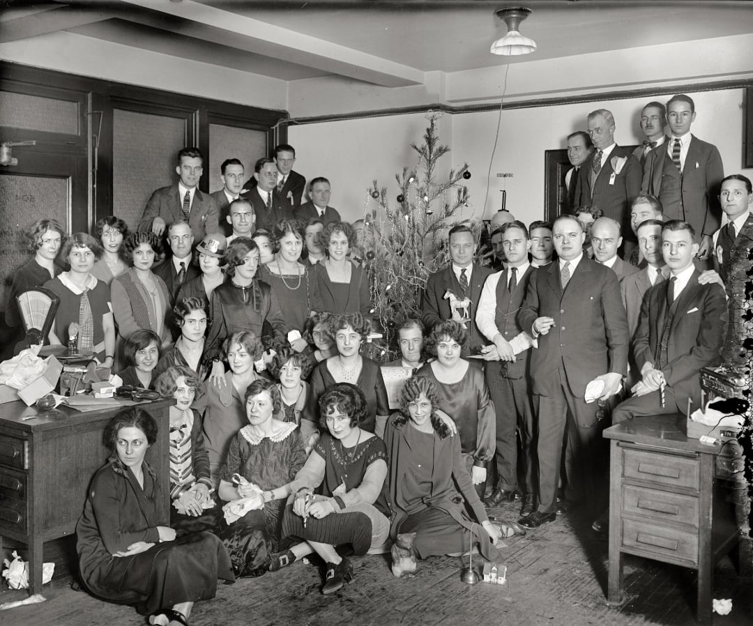 A large group of men and women in 1920s attire gather around a decorated Christmas tree in an office, posing for a holiday photo with desks and paperwork visible in the background.