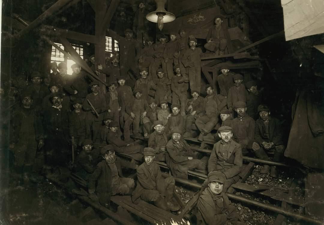 A large group of young boys wearing work clothes and hats pose inside a dark, industrial building, possibly a factory or mine, surrounded by machinery and wooden beams.