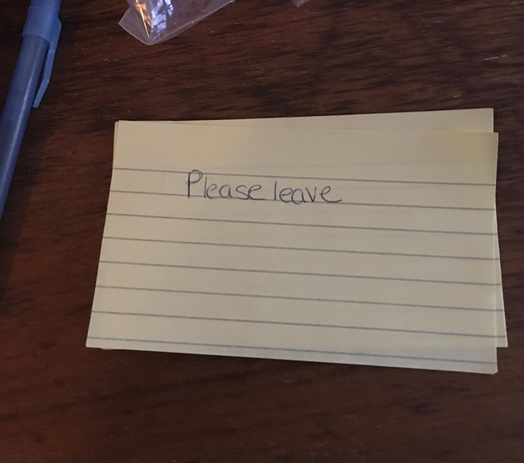 A yellow lined notecard with the handwritten words “Please leave” sits on a dark wooden surface next to a blue pen.