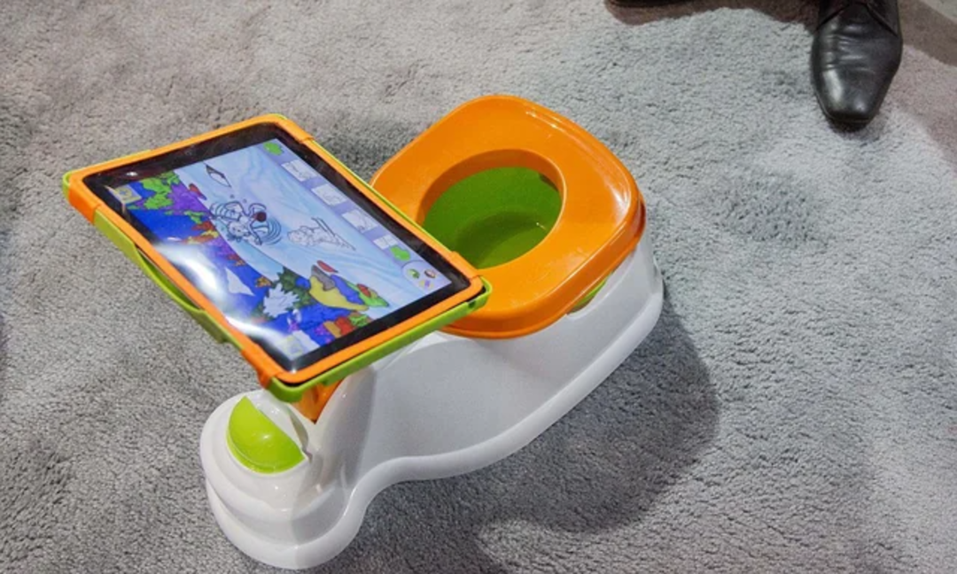 A child's potty training seat with an orange trim and a built-in tablet holder displaying a colorful cartoon, placed on a grey carpeted floor. An adult's shoe is visible in the background.