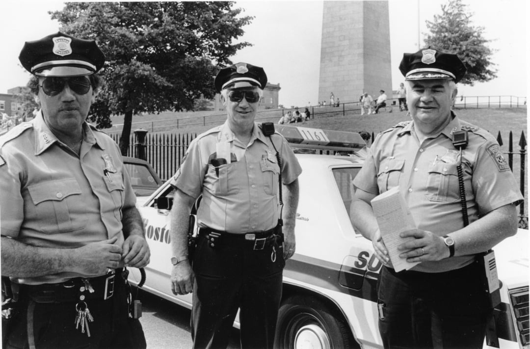 Three uniformed police officers stand in front of a Boston police car, smiling. A tall monument and trees are visible in the background, with people sitting on grassy steps nearby.