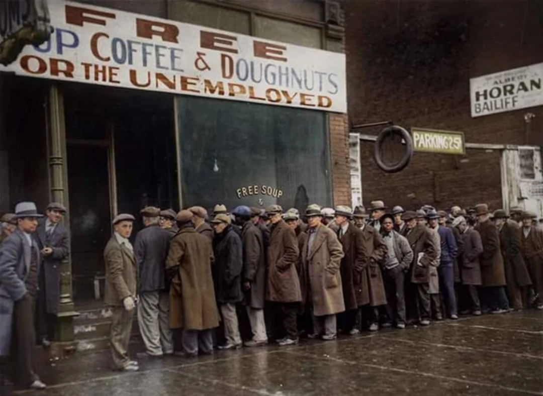 A long line of men in coats and hats wait outside a building with a sign reading "Free Coffee & Doughnuts for the Unemployed." The street is wet, and the scene appears to be from the early 20th century.