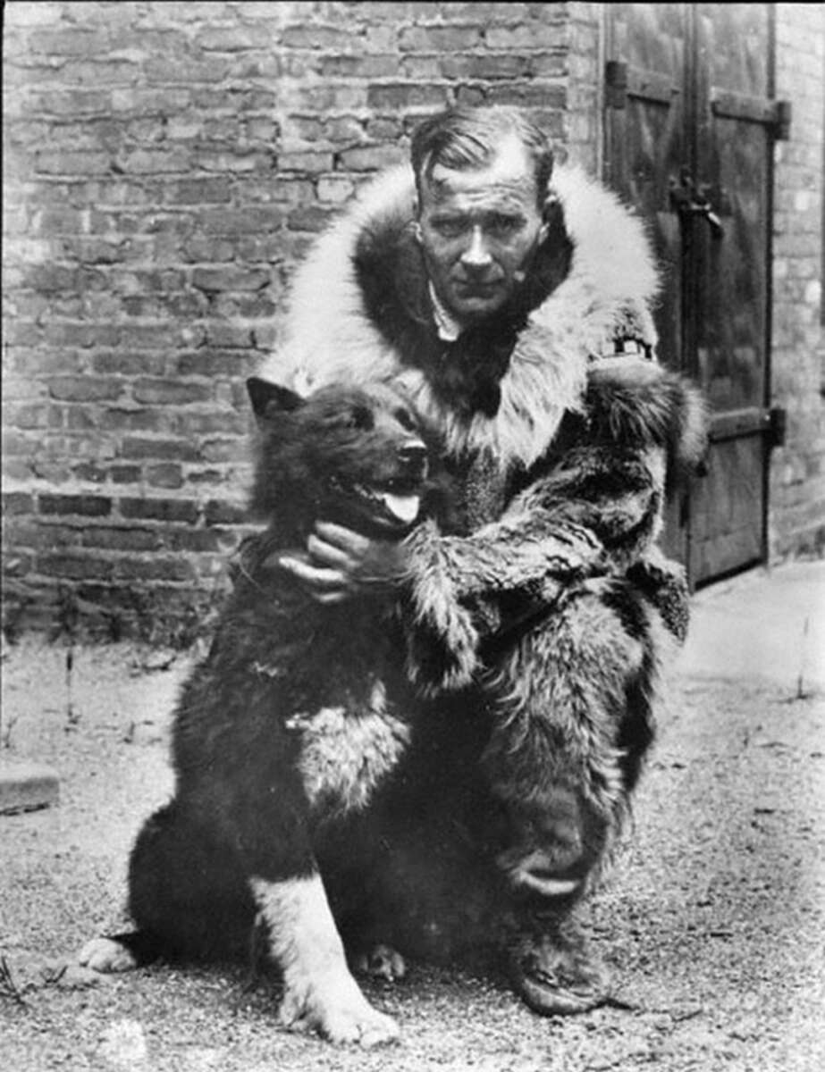 A man in a fur coat kneels beside a large, fluffy dog, holding it gently; both are outdoors near a brick wall and wooden doors.