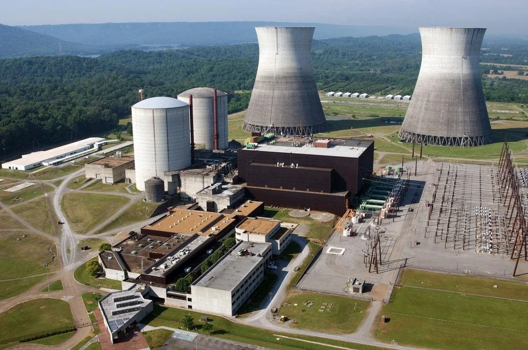 Aerial view of a power plant with two large cooling towers, cylindrical storage tanks, various buildings, and electrical infrastructure surrounded by green fields and trees.