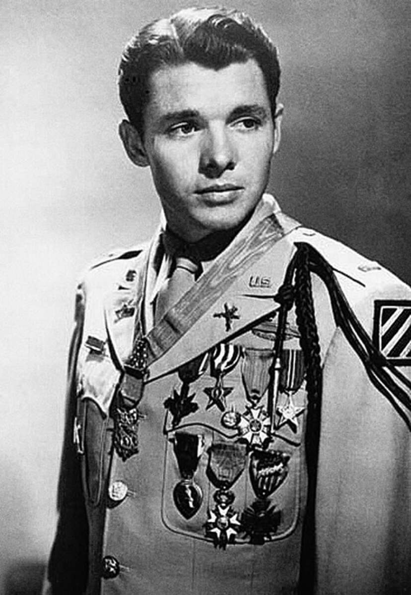 A young man in a decorated military uniform stands facing slightly left, looking away from the camera. His uniform is adorned with numerous medals and badges, and the background is plain and light colored.