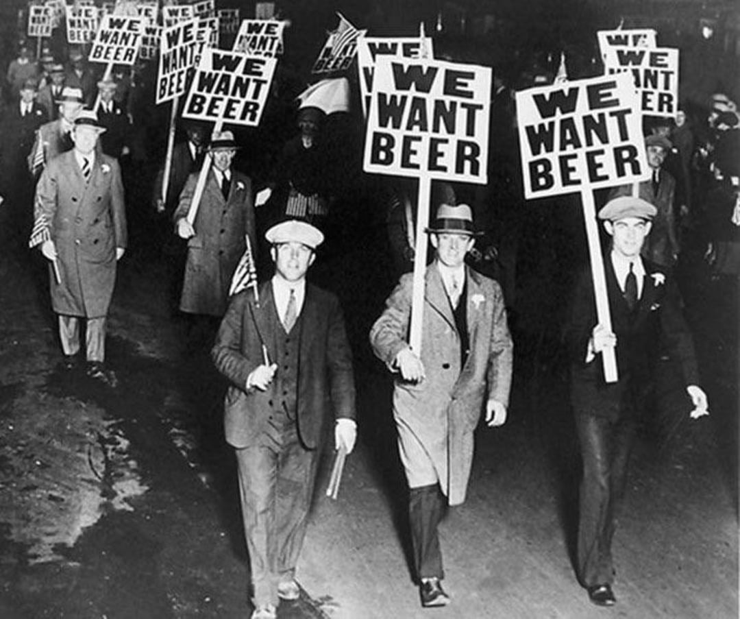 A black-and-white photo showing a group of men in suits and hats marching in protest, holding large signs that read "WE WANT BEER." The scene appears to be from the Prohibition era.