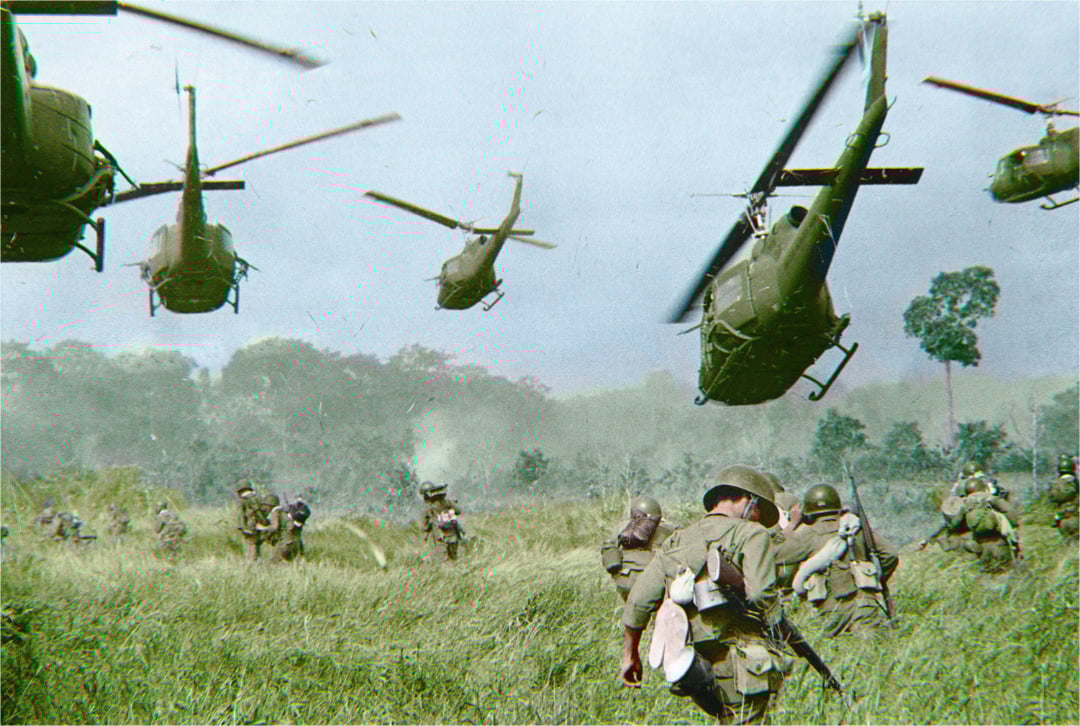 Helicopters hover above a grassy field as soldiers, some crouched and some standing, move through the tall grass during a military operation. Smoke rises in the background and trees are visible in the distance.