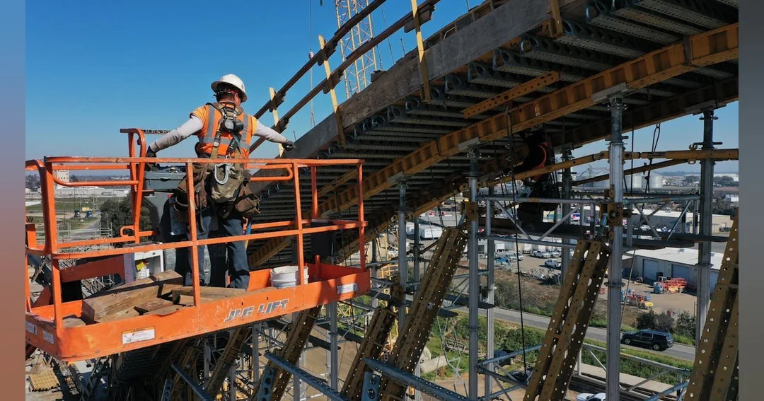 A construction worker in safety gear stands on an orange lift, elevated beside a large steel and wooden bridge structure under construction, with scaffolding and an industrial area visible below.