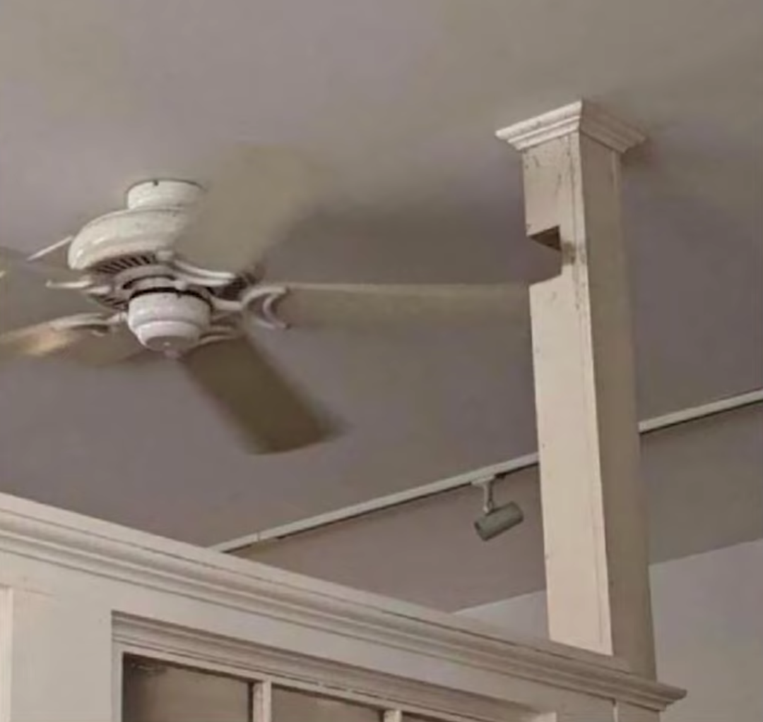 A ceiling fan is spinning with one of its blades wedged into a vertical support beam, showing poor placement and resulting in the fan being unable to rotate freely.
