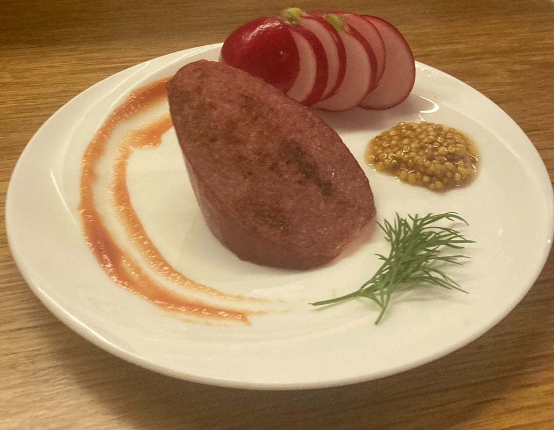 A white plate with a slice of cooked meat, thinly sliced radishes, a dollop of whole grain mustard, a sprig of dill, and a swipe of reddish sauce on a wooden table.