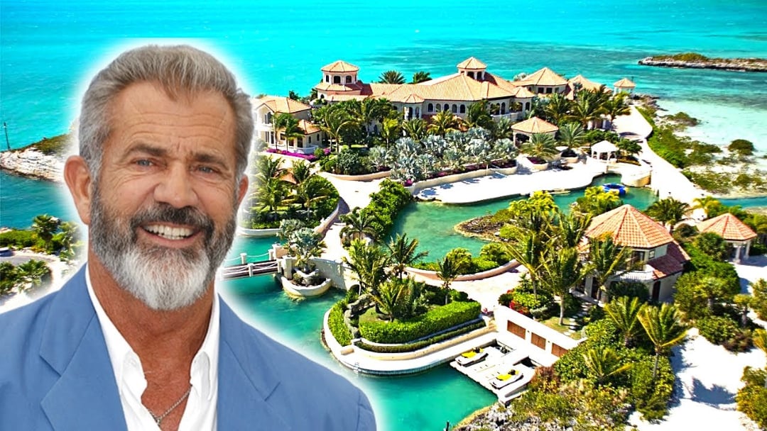 A smiling bearded man in a light blue jacket is shown in front of a luxurious mansion surrounded by turquoise water, palm trees, and lush greenery on a tropical island.