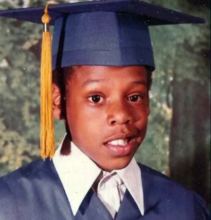A young boy wearing a blue graduation cap and gown with a yellow tassel, smiling slightly at the camera. The background is blurred with green and brown tones.