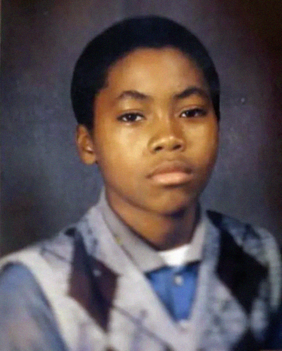 A young boy with short hair wears a collared shirt and a patterned sweater vest, looking directly at the camera with a neutral expression, against a plain studio background.