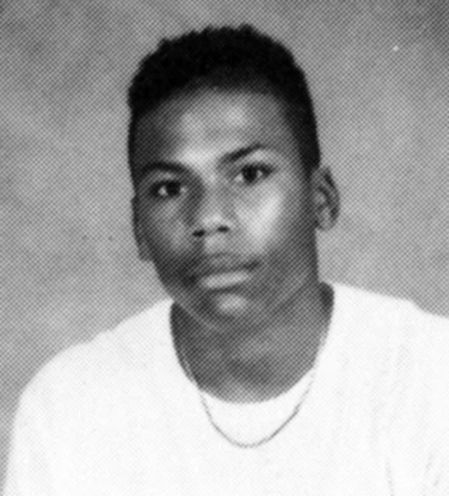 Black and white portrait of a young person with short curly hair, wearing a light-colored shirt and a thin chain necklace, looking directly at the camera with a neutral expression against a plain backdrop.