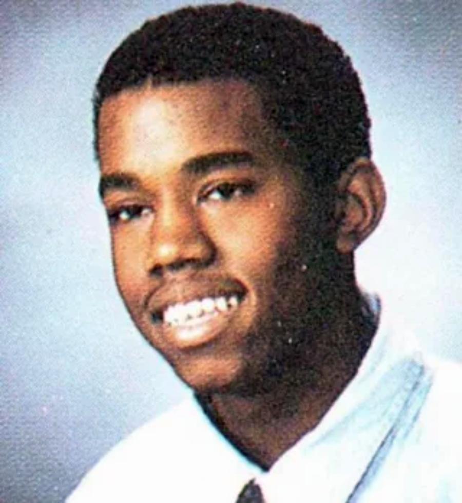 A young Black male smiles in a school portrait, wearing a white collared shirt and a dark tie, against a blue-gray background.