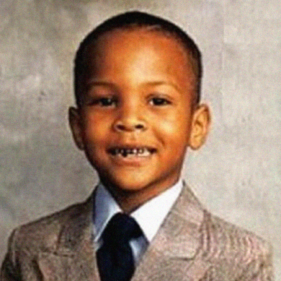 A young boy smiles at the camera, wearing a light-colored suit jacket, white shirt, and dark tie, with a simple gray background behind him.