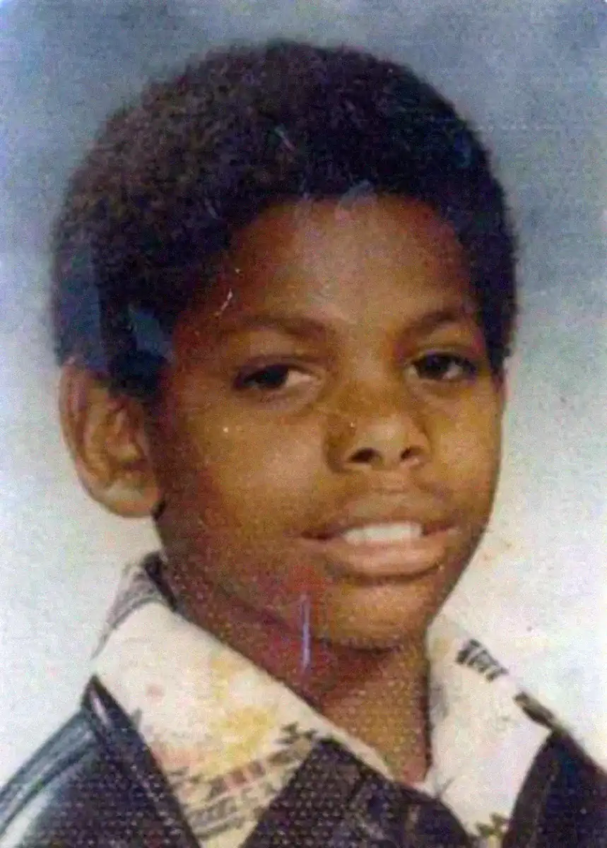 A young boy with short curly hair smiles slightly at the camera. He is wearing a patterned collared shirt under a dark jacket, and the background is a soft, neutral gradient. The photo appears to be a school portrait.