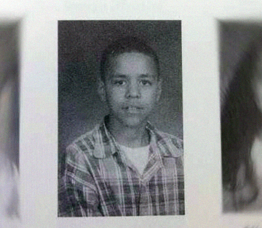 Black-and-white yearbook photo of a young boy wearing a plaid shirt over a white t-shirt, facing the camera with a neutral expression.