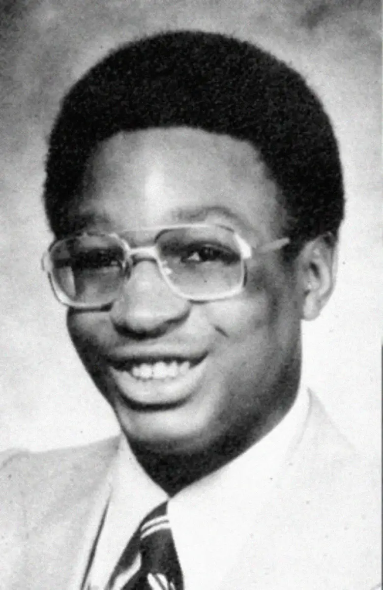 Black-and-white yearbook photo of a young man with short hair, wearing large glasses, a light-colored suit, a white shirt, and a striped tie, smiling at the camera against a neutral background.