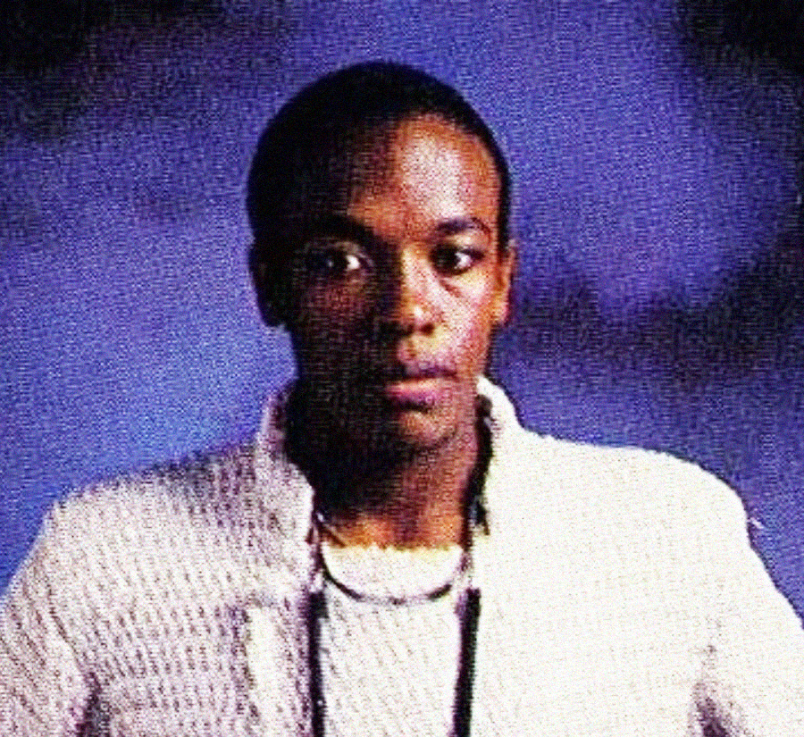 A person with short hair, wearing a light-colored textured jacket and a necklace, stands against a softly lit purple and black background, looking slightly off-camera with a neutral expression.