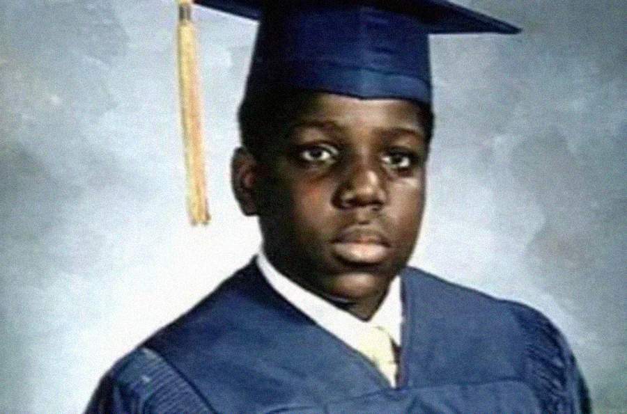 A young boy wearing a navy blue graduation cap and gown poses for a formal school portrait against a neutral background. He has a serious expression and a yellow tassel hangs from his cap.
