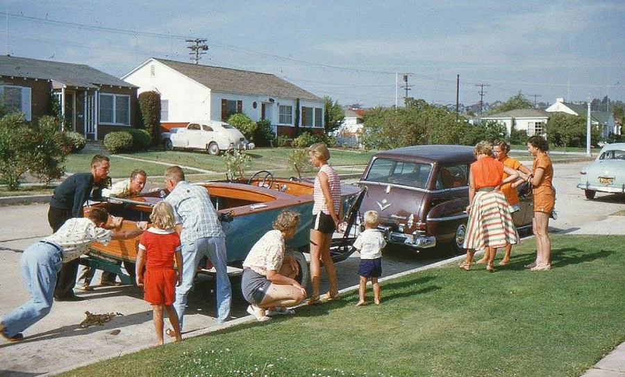 A group of adults and children gather around a wooden boat on a trailer next to a vintage car in a suburban neighborhood, with single-story houses and lawns in the background. The scene appears lively and cheerful.