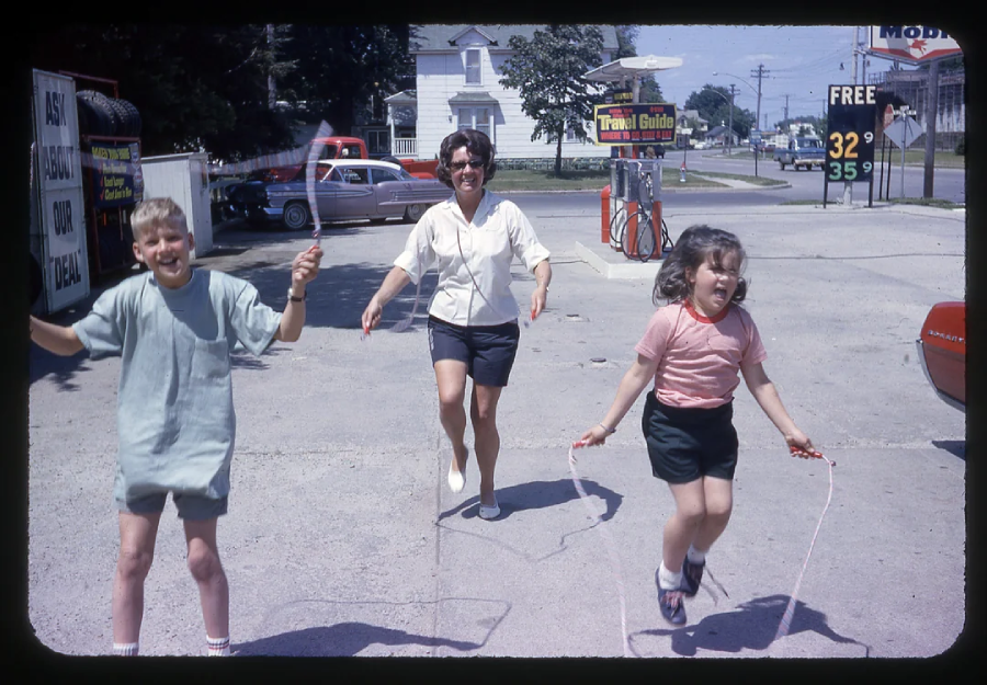 Three people, two children and an adult, are joyfully jumping rope in a gas station parking lot on a sunny day. A gas price sign and vintage cars are visible in the background.