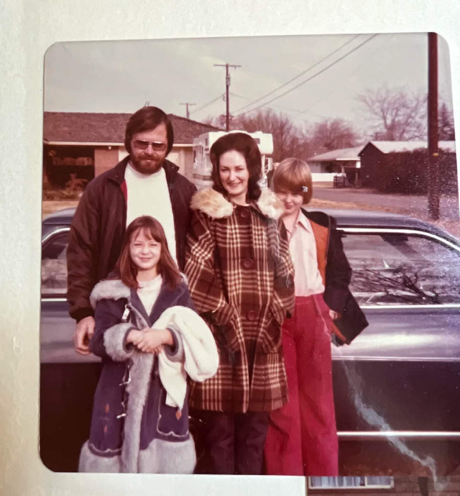 A man, woman, and two young girls stand smiling in front of a vintage car on a suburban street. The adults wear coats and the girls wear winter jackets, suggesting it is a cool day.