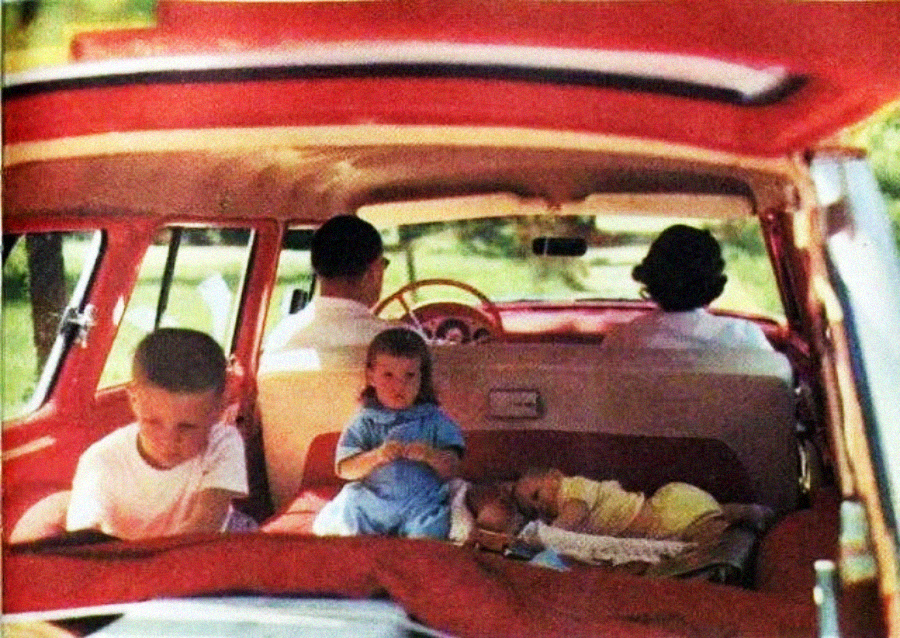 A vintage photo shows two adults in the front seats and three children in the back of a red car. One child is sitting and looking down, another sits in the middle, and a baby lies on a blanket. The scene looks candid and relaxed.