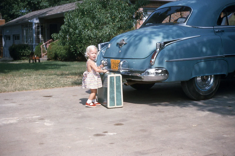 A young child in a striped dress stands on a driveway next to a blue vintage car, holding a green and white suitcase. A house and trees are visible in the background on a sunny day.