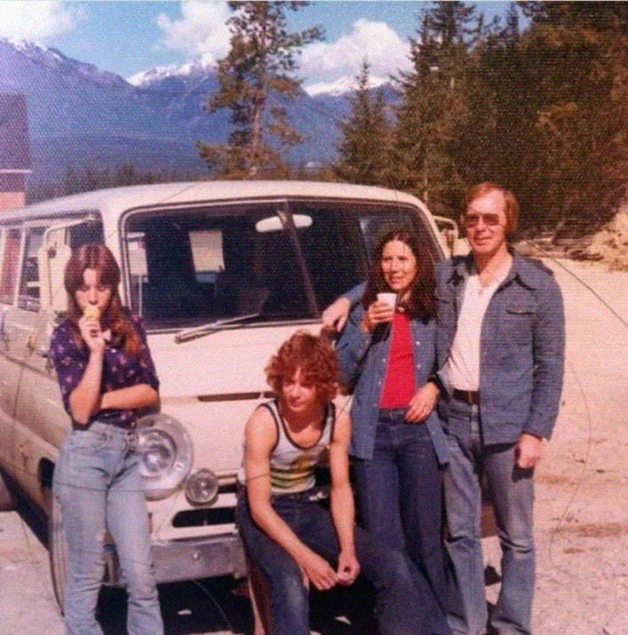 Four people pose in front of a beige vintage van parked outdoors, with mountains and trees in the background. Three stand while one person sits on the van’s bumper. The group appears relaxed and casual.