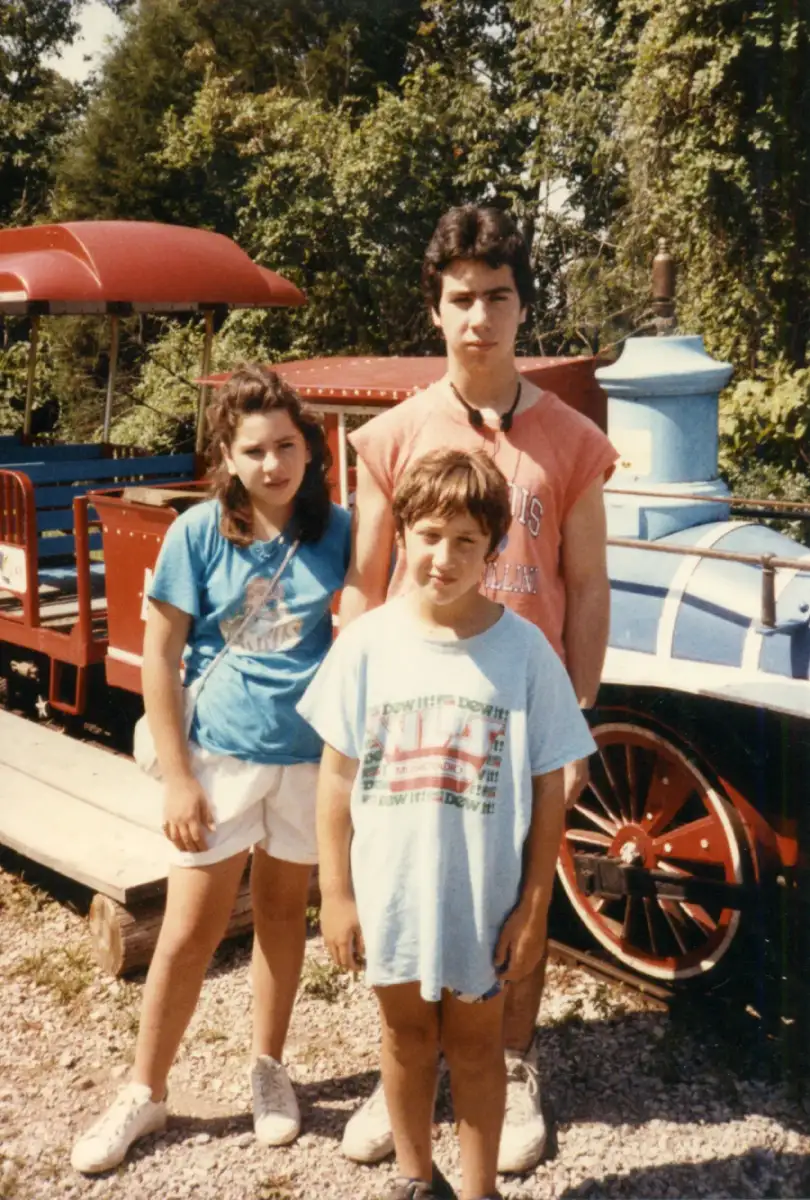 Three children stand in front of a small red and blue train outdoors on a sunny day, surrounded by trees. They are wearing casual t-shirts and shorts, and all look toward the camera with neutral expressions.
