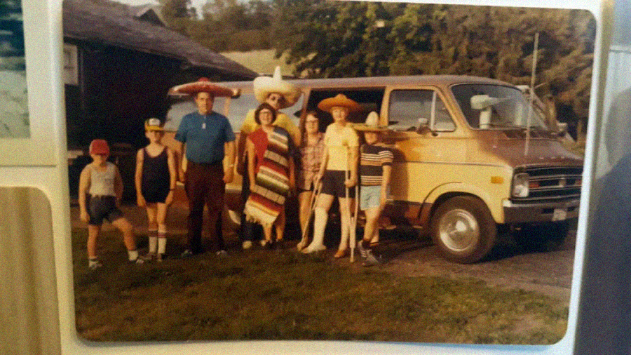 A group of eight people, including children and adults, stand in front of a tan van. Some wear sombreros and colorful ponchos, suggesting a festive or vacation setting. Trees and buildings are visible in the background.