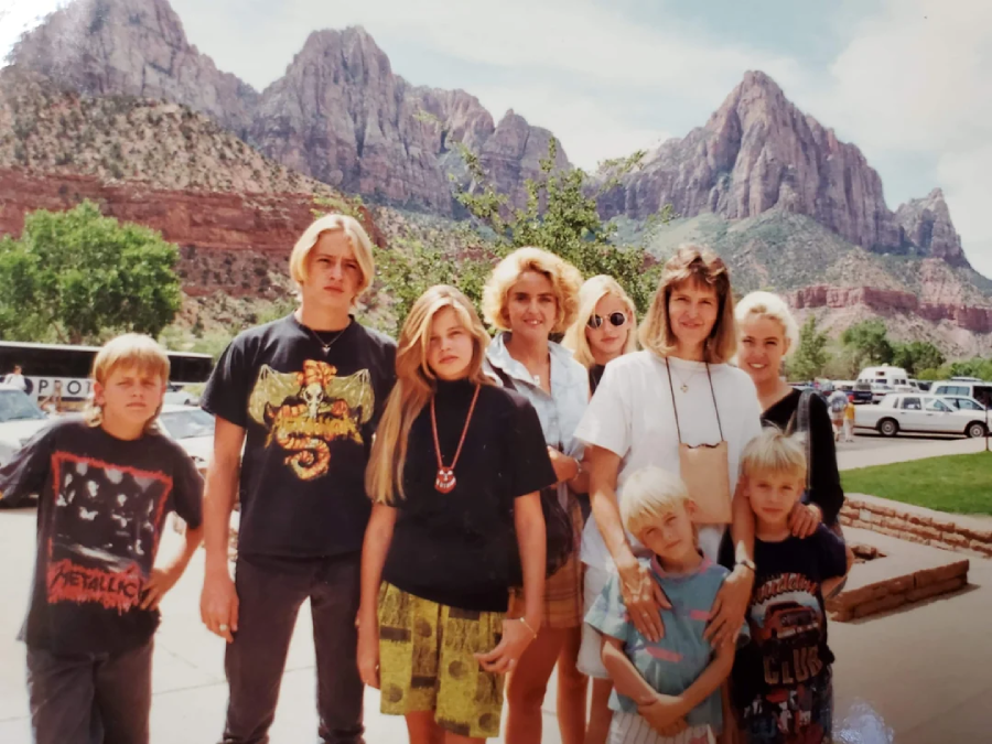 A group of nine people, including children and adults, pose for a photo outdoors in front of a scenic backdrop of rocky mountains and greenery on a sunny day. Some are smiling, and several wear casual clothing with graphic prints.