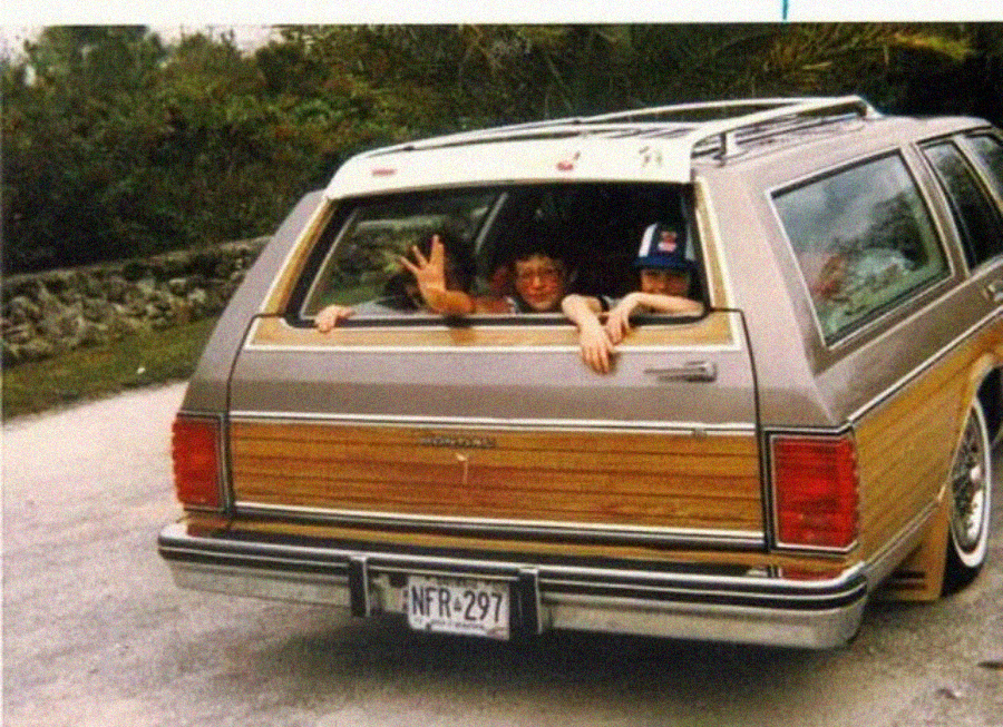 Two children smile and wave from the open rear window of a brown station wagon with wood paneling, parked on a road next to trees. The car has a roof rack and a visible license plate.