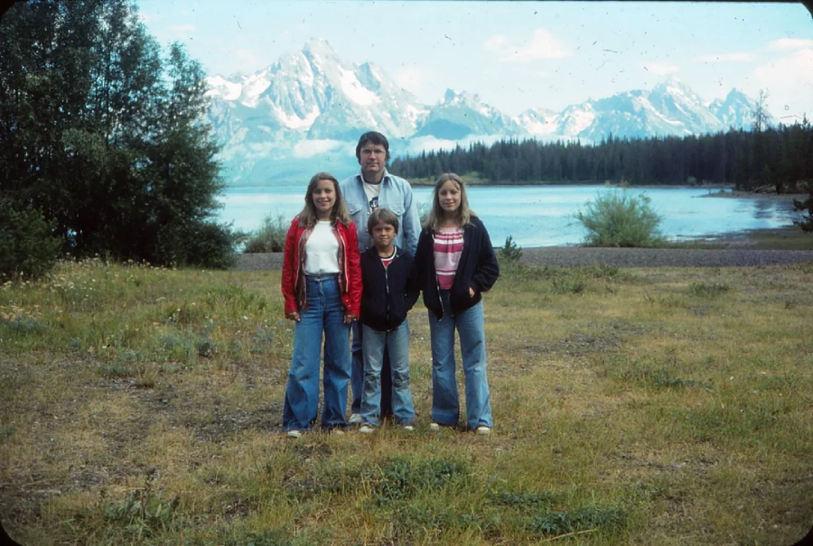 A man and three children stand on grassy ground near a lake, with tall trees and snow-capped mountains in the background. The sky is clear and everyone is wearing jackets and jeans.