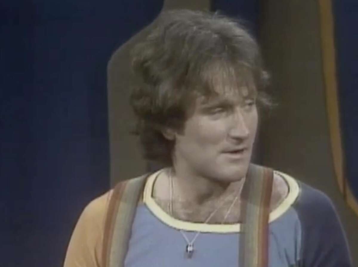 A person with wavy brown hair wears a blue shirt with multicolored suspenders and a necklace, looking to the side against a blurred background.