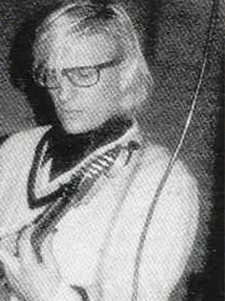 Black and white photo of a person with light-colored, short hair wearing glasses and a sweater. The person is looking slightly downward and to the side, with a blurred background and a visible cable in the foreground.